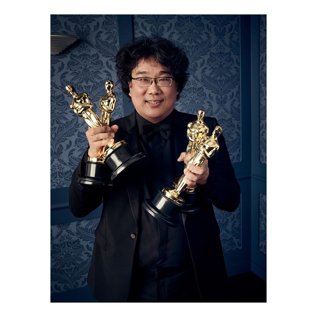 Man in tuxedo holding three golden Oscar awards in studio setting against a blue floral backdrop, celebrating achievement in entertainment.