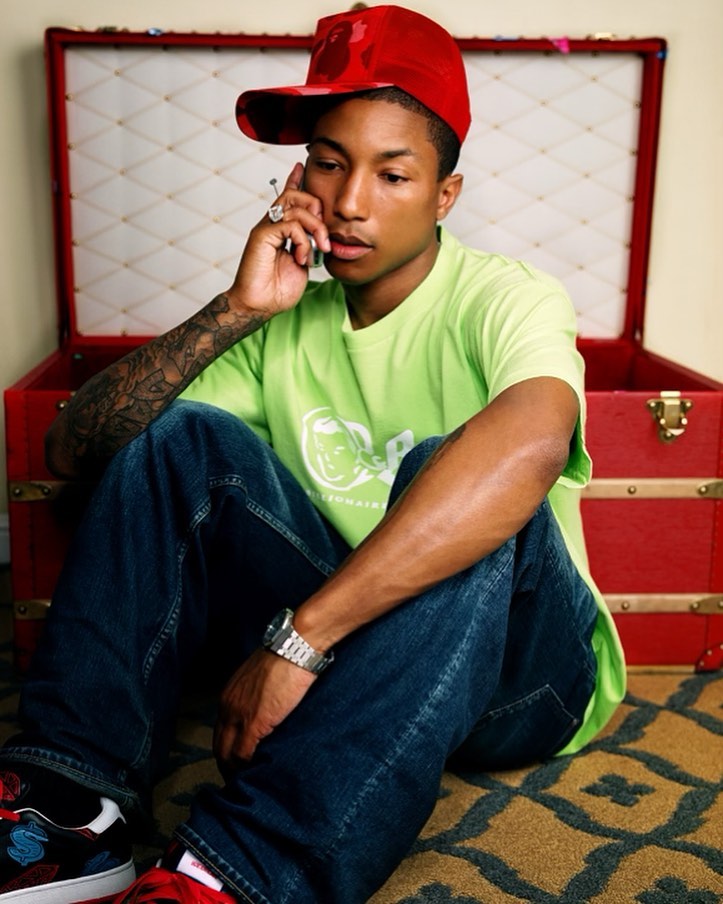 An adult male seated indoors, wearing a red cap and lime green shirt, is talking on a cellphone. He is posed in front of a red storage chest.