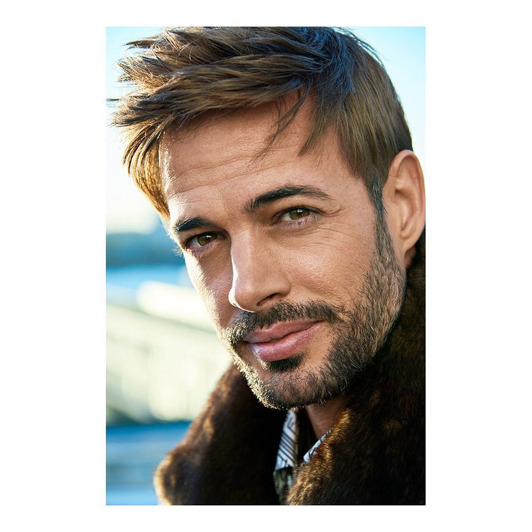 Portrait of a handsome adult male with dark hair and stubble wearing a fur-collared coat against a light blue outdoor background.