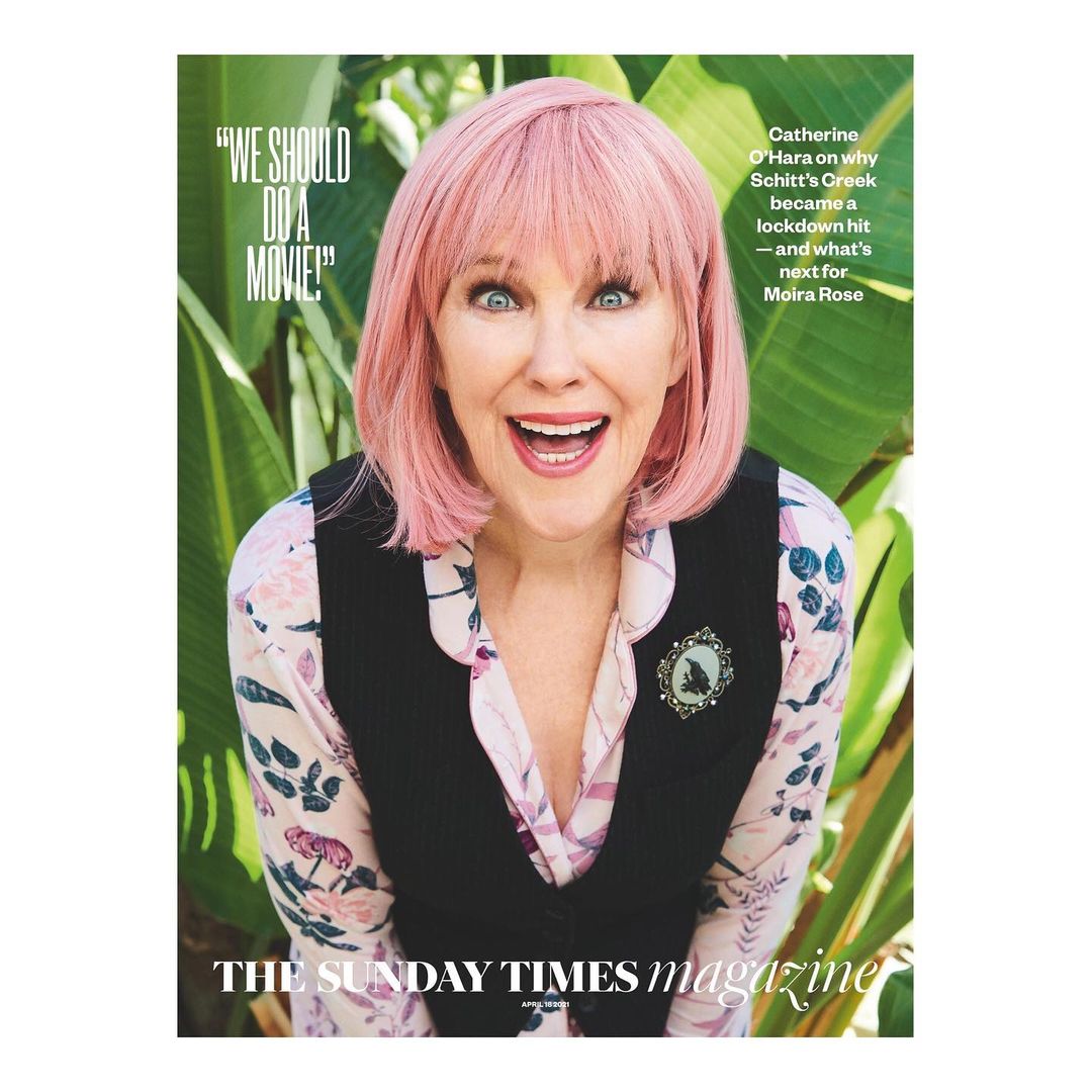 Catherine O'Hara, pink hair, smiles on magazine cover. She wears a floral blouse with black vest in an outdoor setting surrounded by plants.