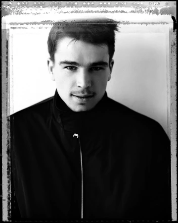 Monochrome portrait of a young man wearing a zipped black jacket against a white background with visible film borders