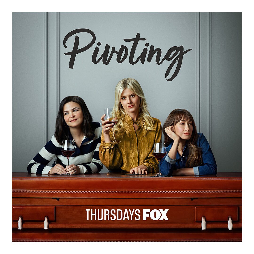 Three women are sitting behind a wooden structure holding wine glasses in front of a grey wall with 'Pivoting' written above them.