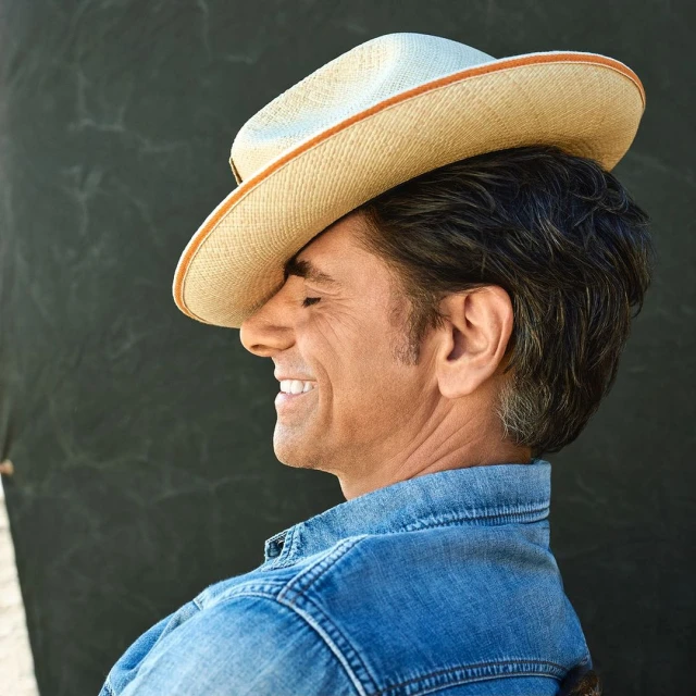 Profile portrait of adult male smiling with a straw cowboy hat on his head, wearing a blue denim jacket against a grey background.