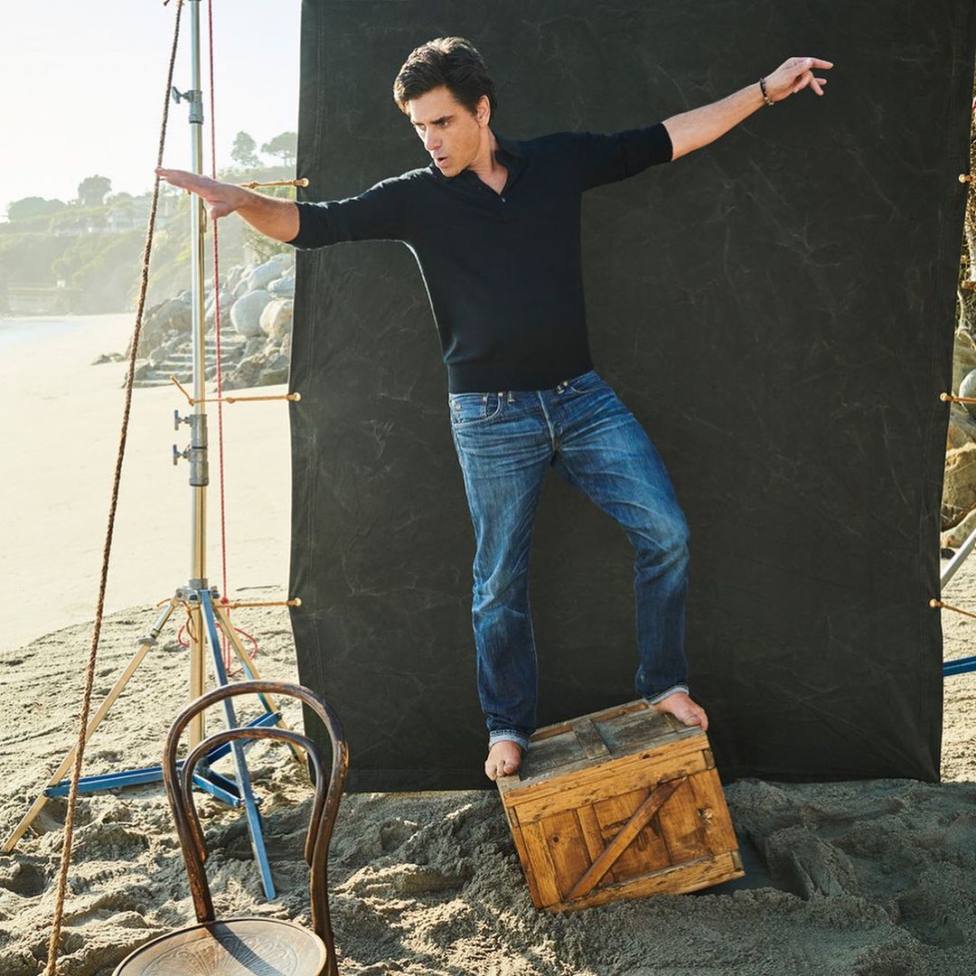 Man in casual clothes balances on a wooden box on a sandy beach with ocean and cliffside views, posing with arms outstretched.