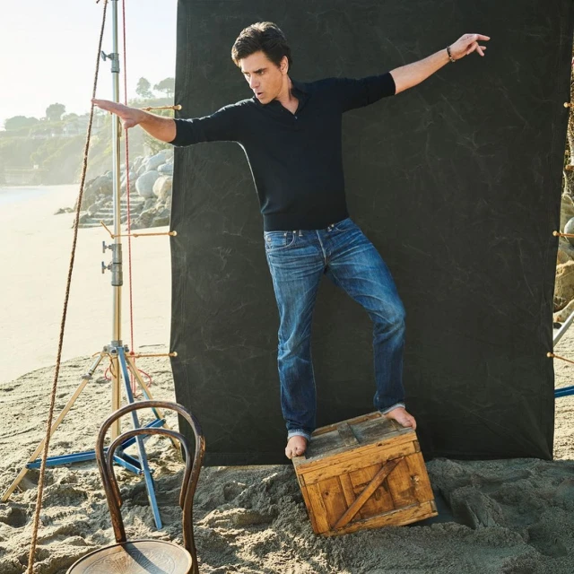 Man in casual clothes balances on a wooden box on a sandy beach with ocean and cliffside views, posing with arms outstretched.