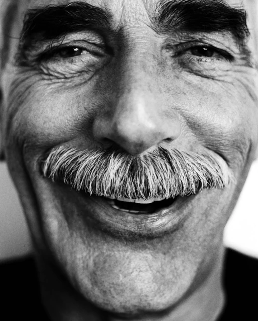 Close-up black and white portrait of a smiling elderly man with a thick, well-groomed mustache. High detail shows skin texture and eyes.