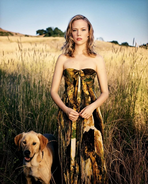 An adult woman in a dress stands in a field of tall grass with a yellow Labrador Retriever dog on a sunny day in a rural environment.