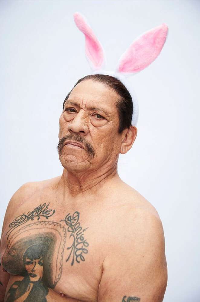 Danny Trejo poses shirtless wearing Easter bunny ears against a white background. He has multiple tattoos on his chest.