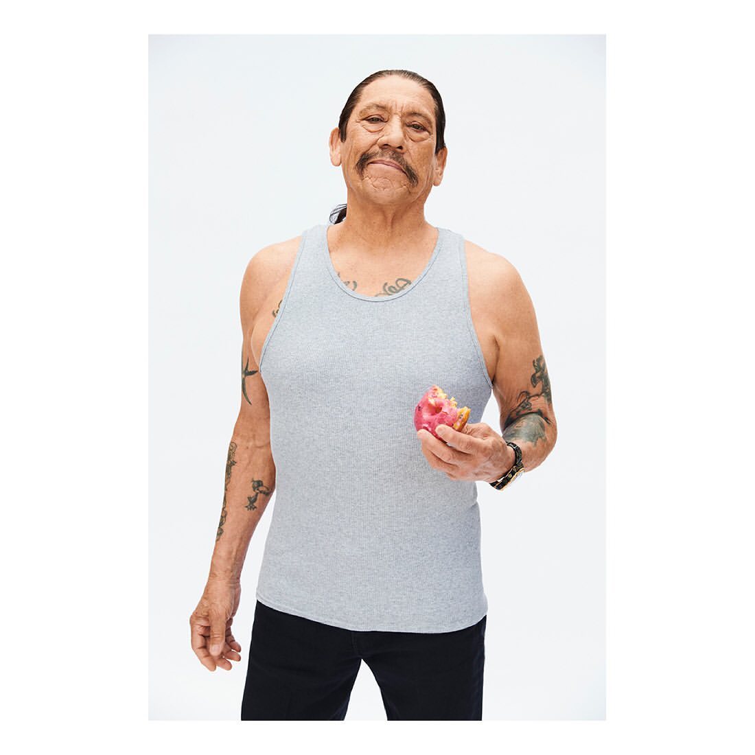 Danny Trejo posing indoors with a bitten red donut. He is wearing a grey tank top and black pants against a white backdrop.