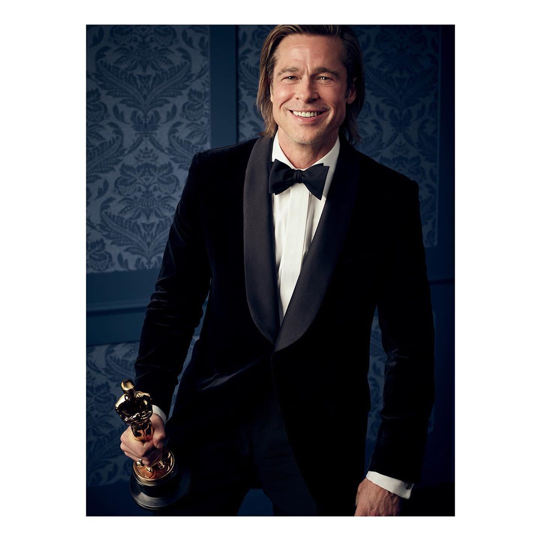 Brad Pitt poses with an Academy Award. He is wearing a black tuxedo and bow tie. The background is a dark blue patterned wallpaper.