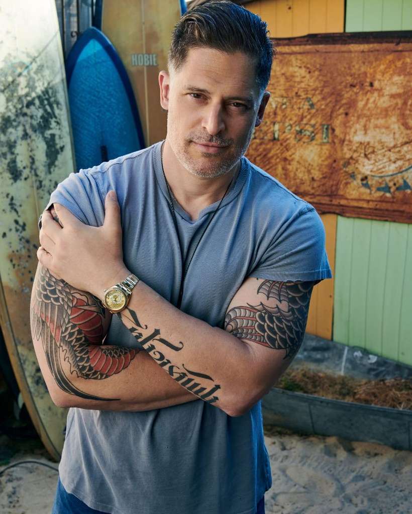 Adult male portrait with arm tattoos standing in front of surfboards and a rustic wall, wearing a gray t-shirt and a gold wristwatch.