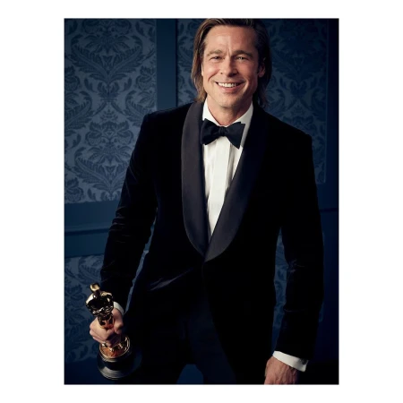 Brad Pitt in a black velvet tuxedo holding an Oscar statuette against a blue patterned wallpaper background