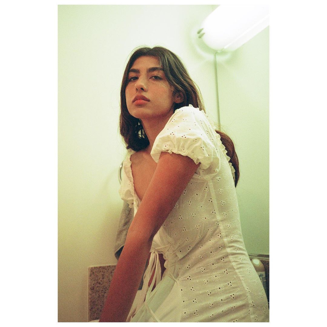 Portrait of a young woman with long brown hair and a white dress looking at the camera indoors with a light and wall in the background.