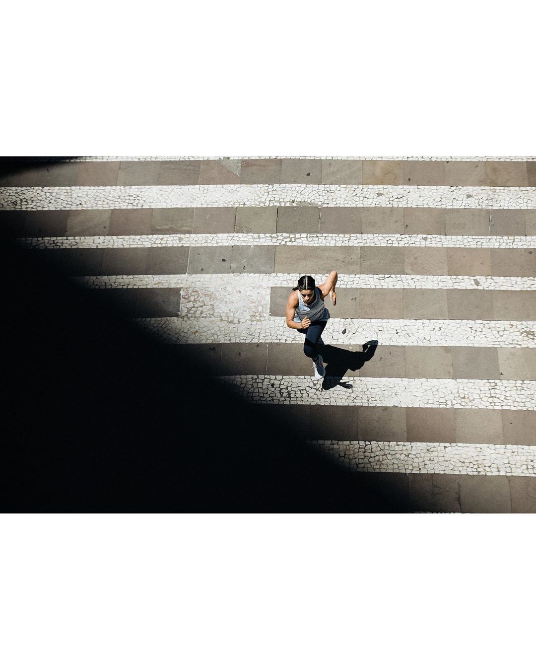 Overhead shot of a woman running on a striped stone pathway in daytime, partially obscured by shadows, creating contrast and movement.
