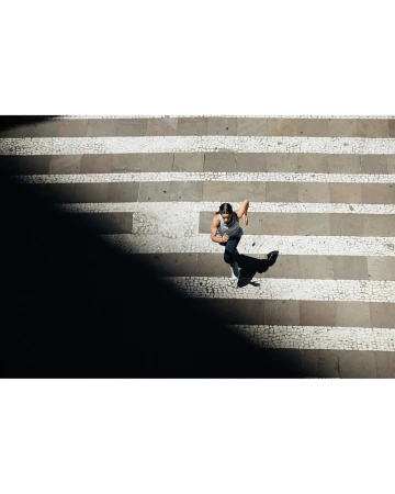 Overhead shot of a woman running on a striped stone pathway in daytime, partially obscured by shadows, creating contrast and movement.