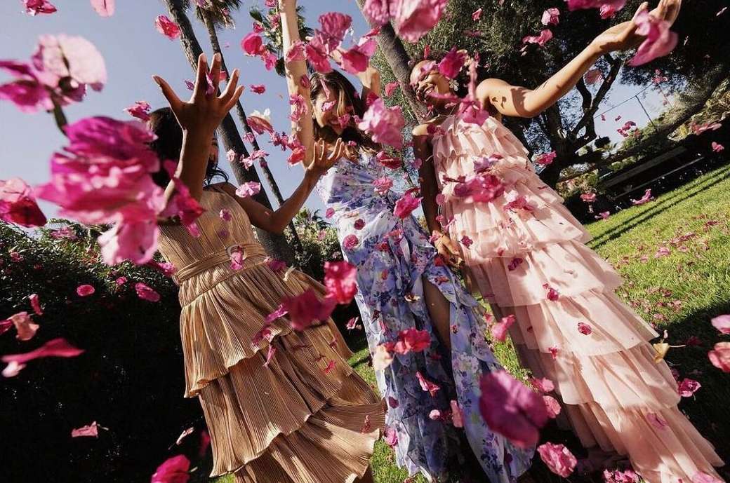 Three women in tiered dresses outdoors, celebrating with pink flower petals falling around them in a summer party atmosphere.