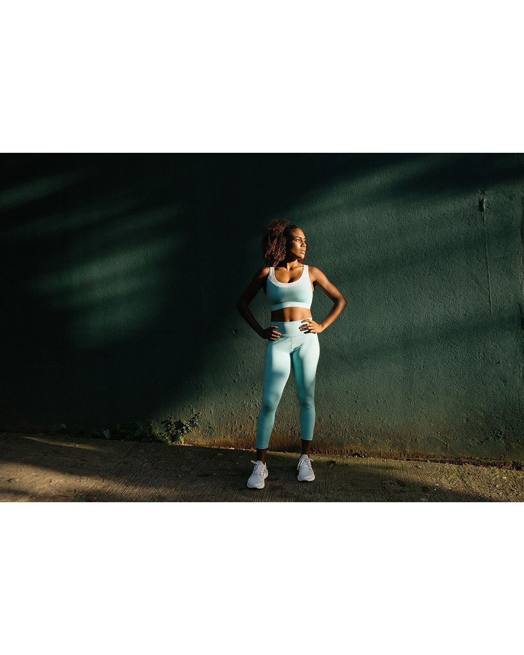 Athletic Black woman in workout gear stands confidently outdoors against a textured wall, bathed in sunlight, showcasing fitness lifestyle.