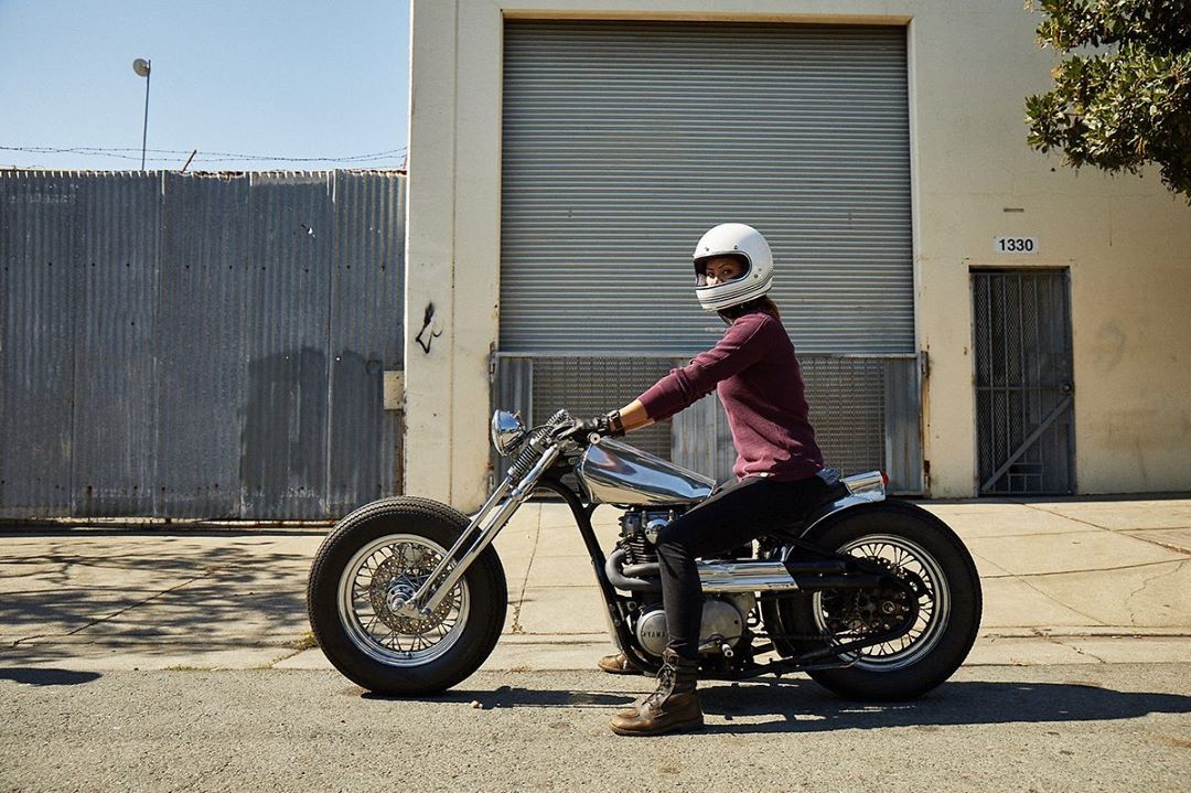 Woman on a custom motorcycle with silver gas tank and white helmet. The vehicle is parked in front of an industrial building.