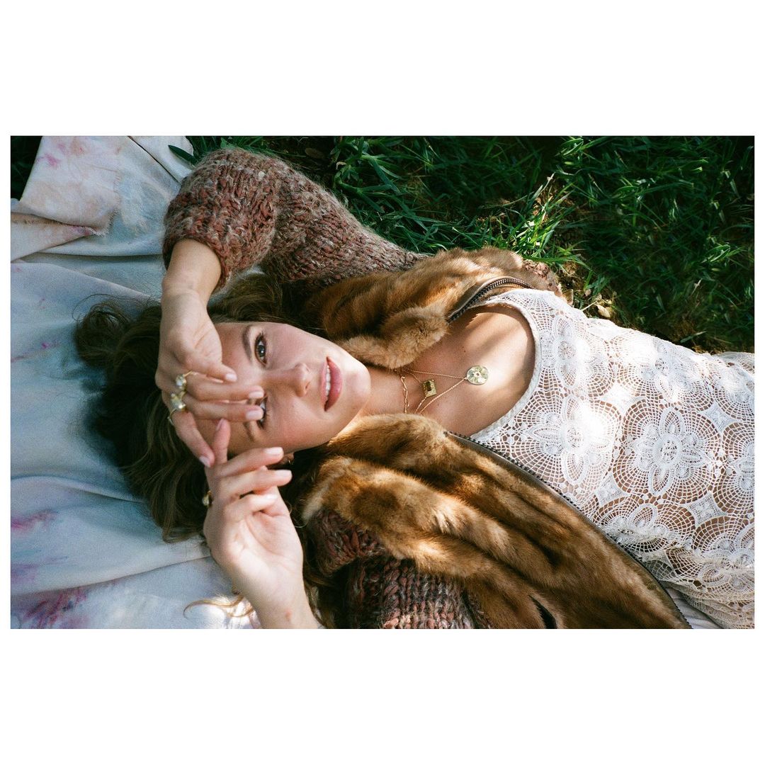 Woman in white lace dress and fur stole lying on a blanket outdoors in green grass, relaxing with sunlight, posing for a portrait.