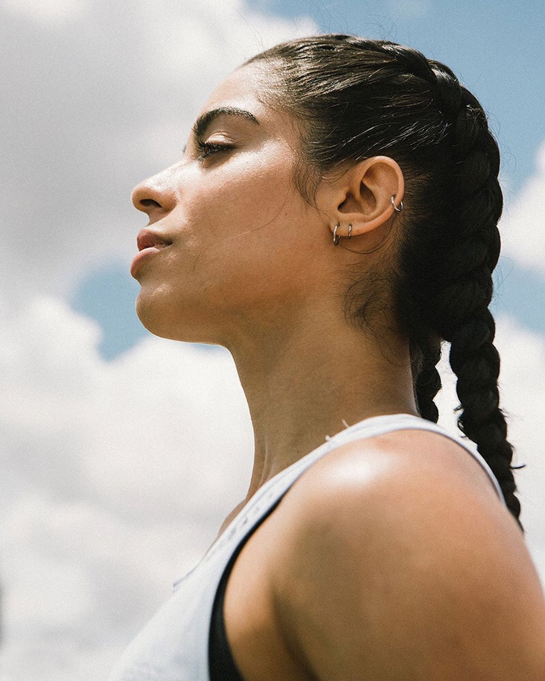 Profile shot of a woman with braided hair wearing a tank top, looking up towards the sky with clouds in the background on a sunny day.