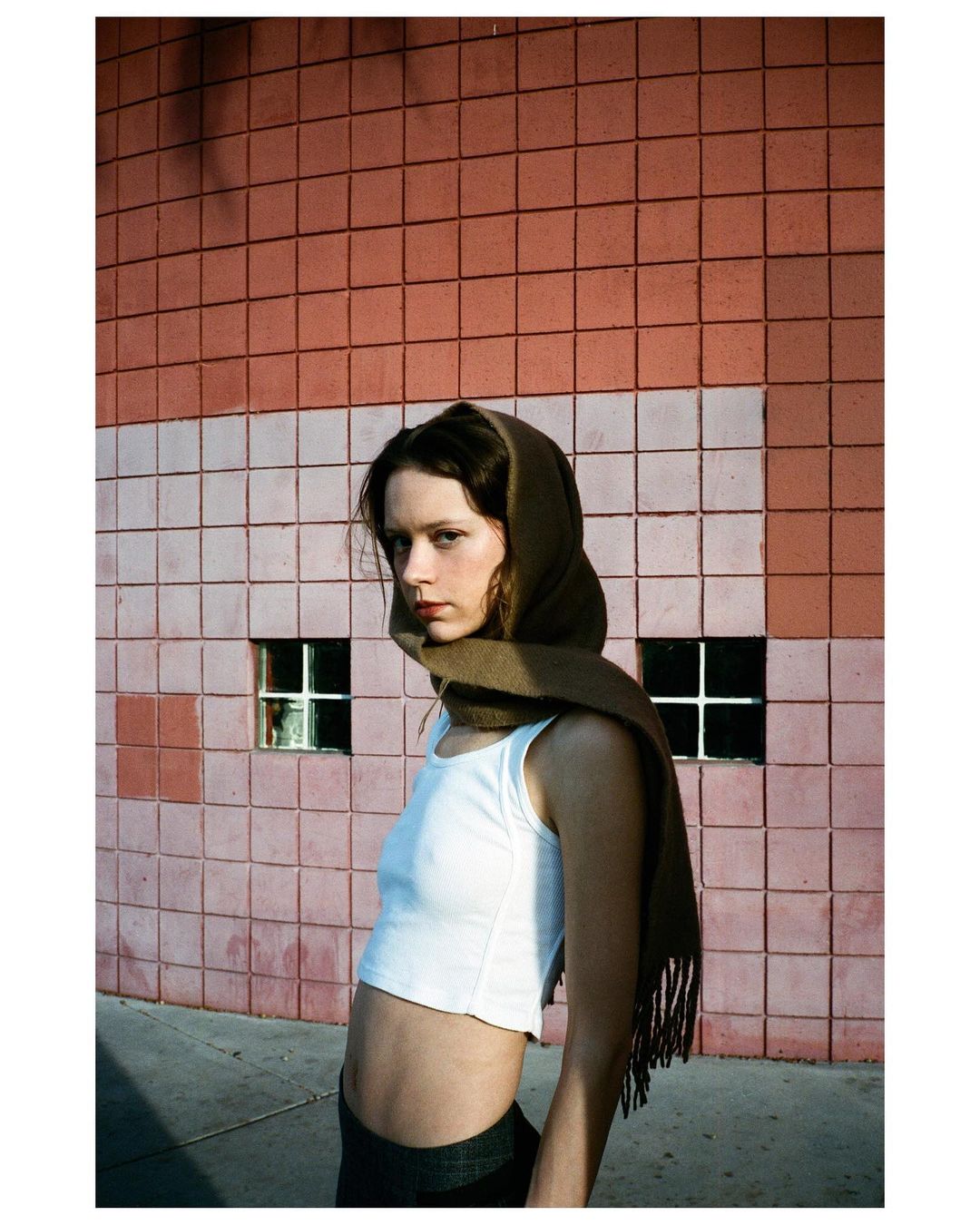 A young woman stands outdoors wearing a white tank top and scarf, posing against a geometric tiled wall with natural, diffused lighting.