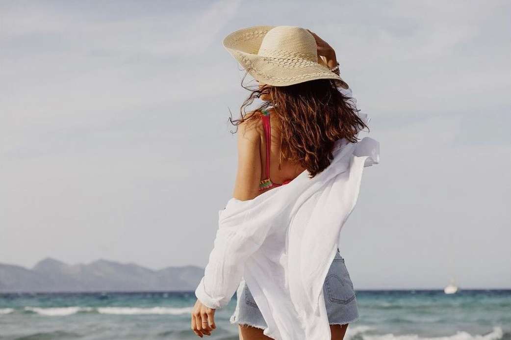 Back view of a woman with brown hair, wearing a straw hat, bikini, and a white shirt at the beach with mountains in the background.