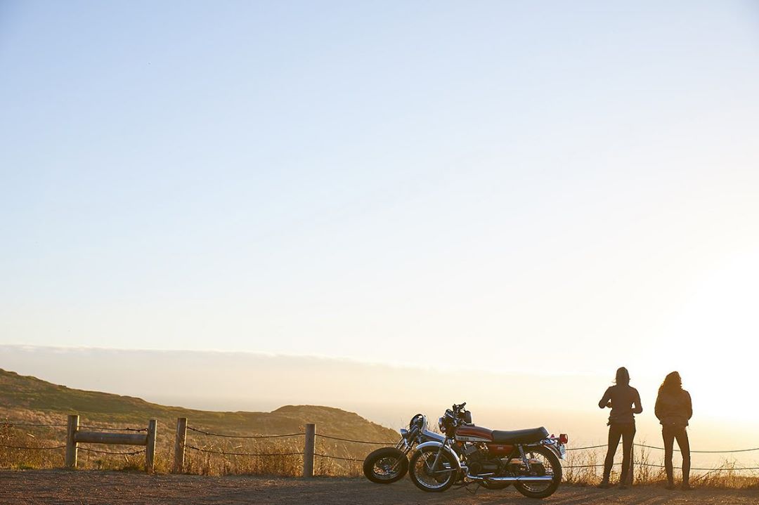 Two people standing near a vintage motorcycle against a coastal sunset backdrop, creating a sense of adventure and freedom.