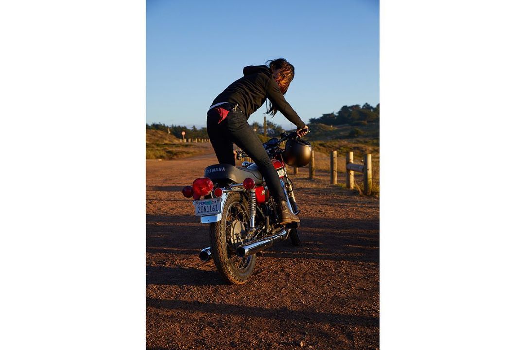 Woman sits on a Yamaha motorcycle with a helmet in her hand on a dirt road with wooden fence posts and distant hills under a clear sky.