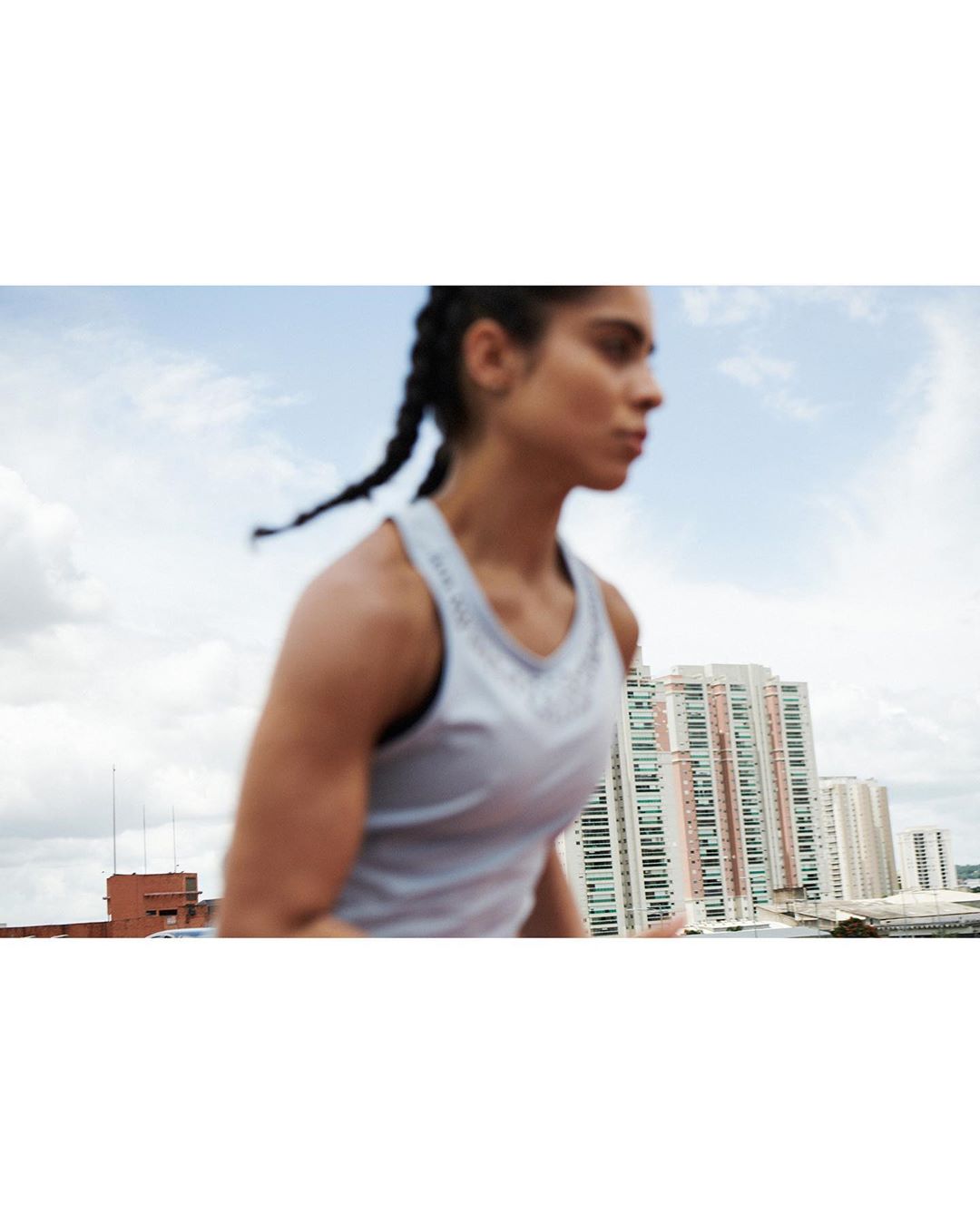Athletic woman in tank top training outdoors in a city with high rise buildings under a cloudy sky, side view, medium shot.