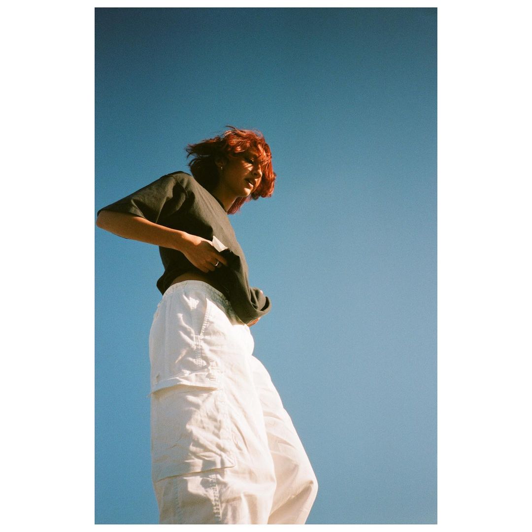 Low angle shot of a young adult woman with red hair, wearing a gray t-shirt and white pants, posing against a clear blue sky.