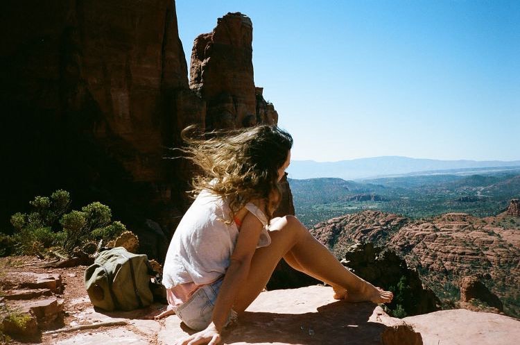 Woman sits on rocky outcrop overlooking a mountain range. The view includes red rock formations, green vegetation, and a blue sky.