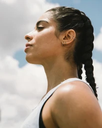 Profile portrait of a woman with braided dark hair wearing a white tank top against a clear sky background