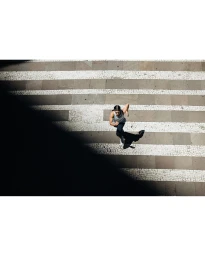 High angle shot of a woman running across a patterned paved street with deep shadow cast across the lower left corner.