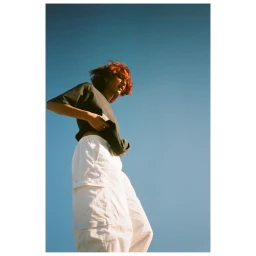 Low-angle shot of a person with short red hair wearing a dark t-shirt and white cargo pants against a clear blue sky background.