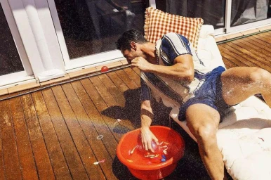 Man in a patterned short-sleeve shirt and blue shorts splashing water from a red basin while seated on a lounger on a wooden deck