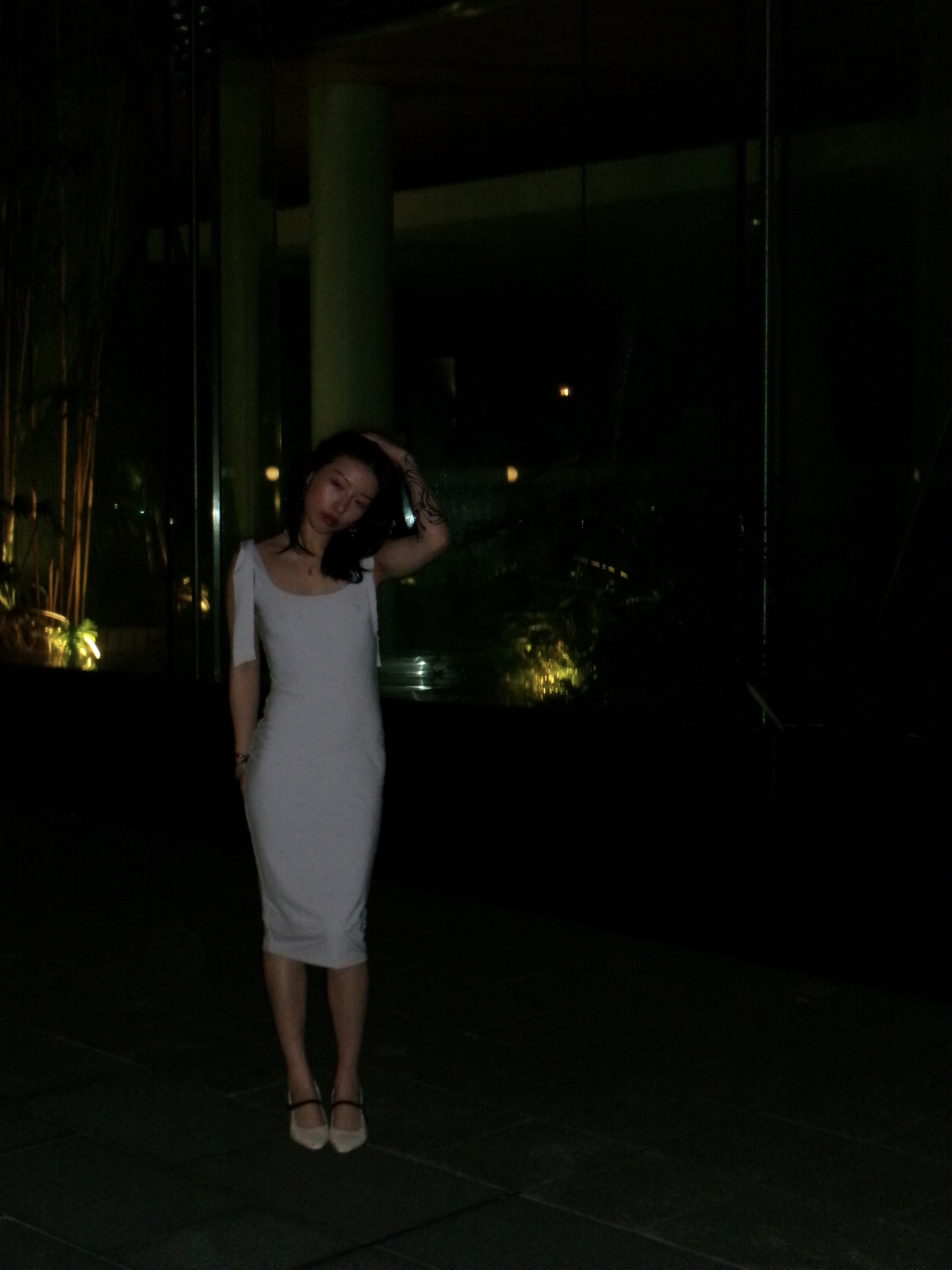 Adult woman with dark hair poses in a grey dress with hand raised to her head in a nighttime setting with visible city lights.