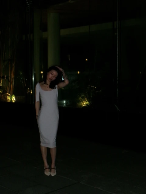 Adult woman with dark hair poses in a grey dress with hand raised to her head in a nighttime setting with visible city lights.