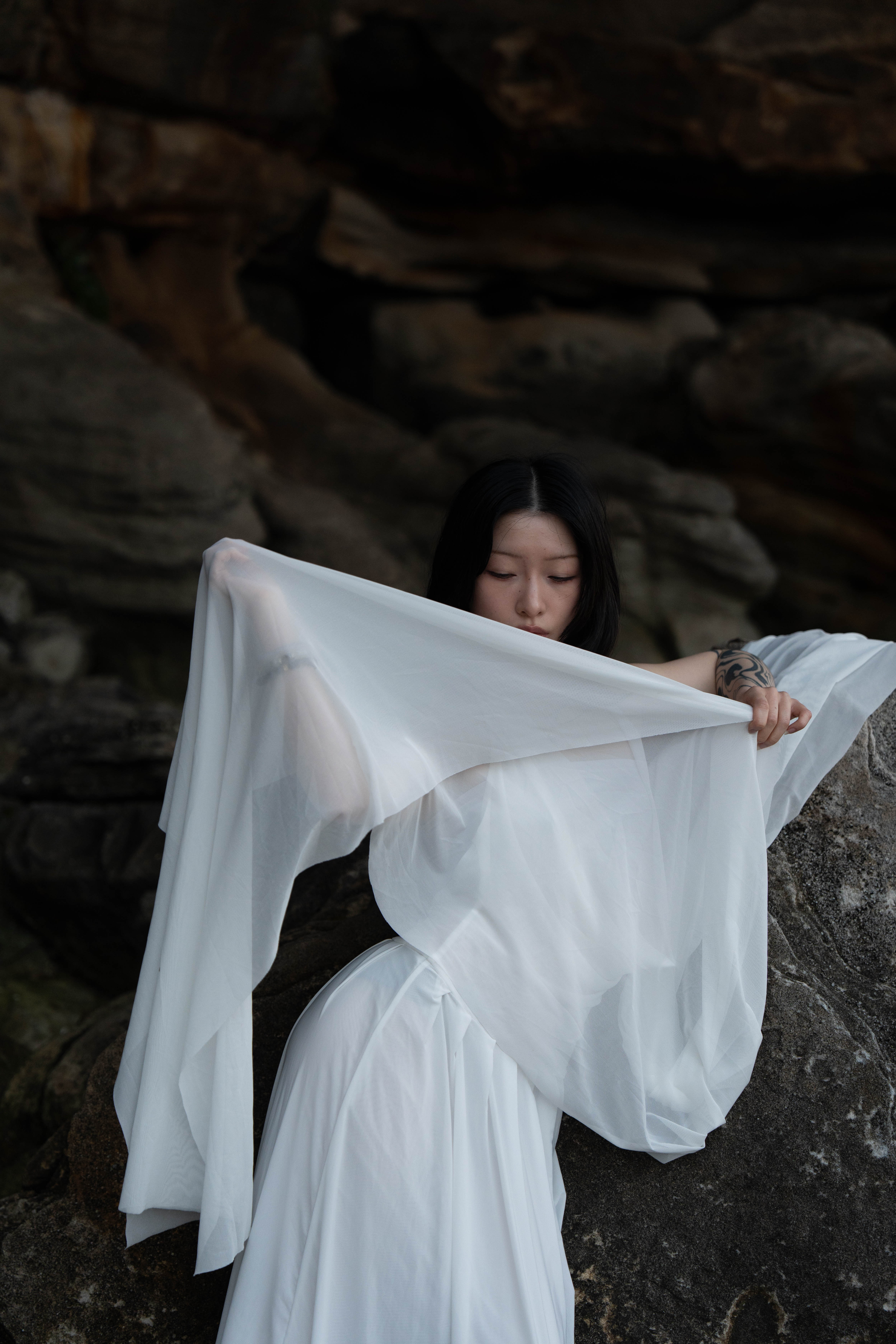 Model Savalia holding sheer white fabric across a draped white dress against dark textured rock formations.