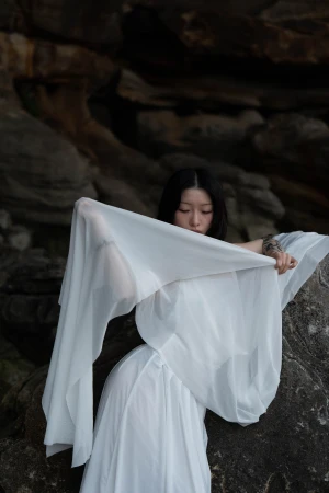 Model Savalia holding sheer white fabric across a draped white dress against dark textured rock formations.