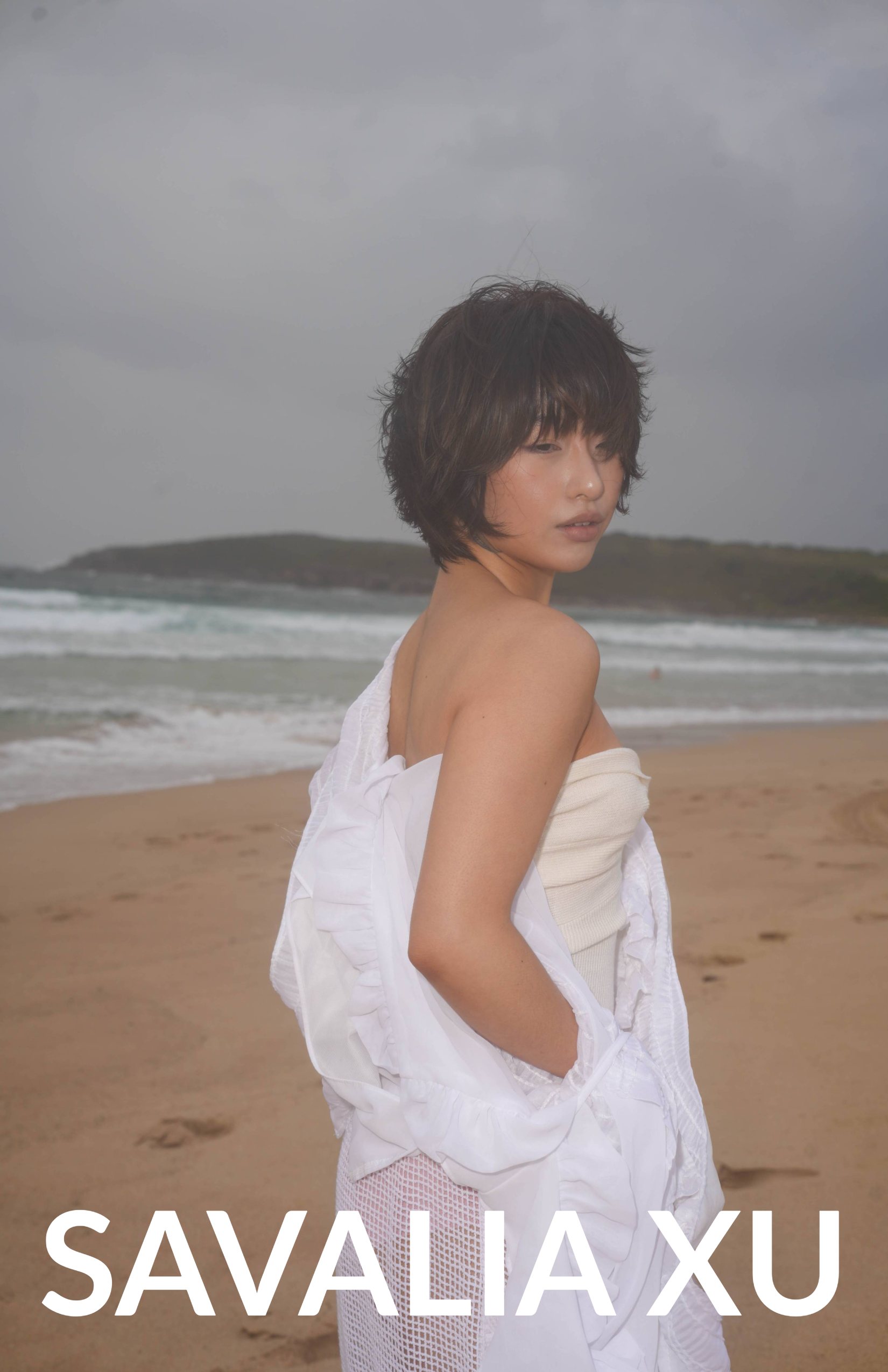 A young woman stands on a beach with ocean waves in the background, wearing a white top and a sheer, draped white fabric.