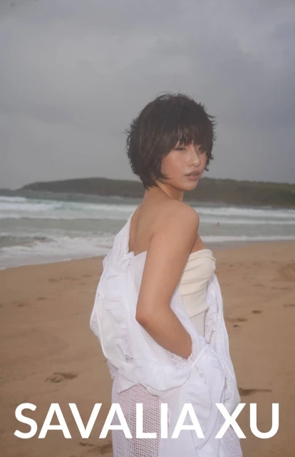A young woman stands on a beach with ocean waves in the background, wearing a white top and a sheer, draped white fabric.