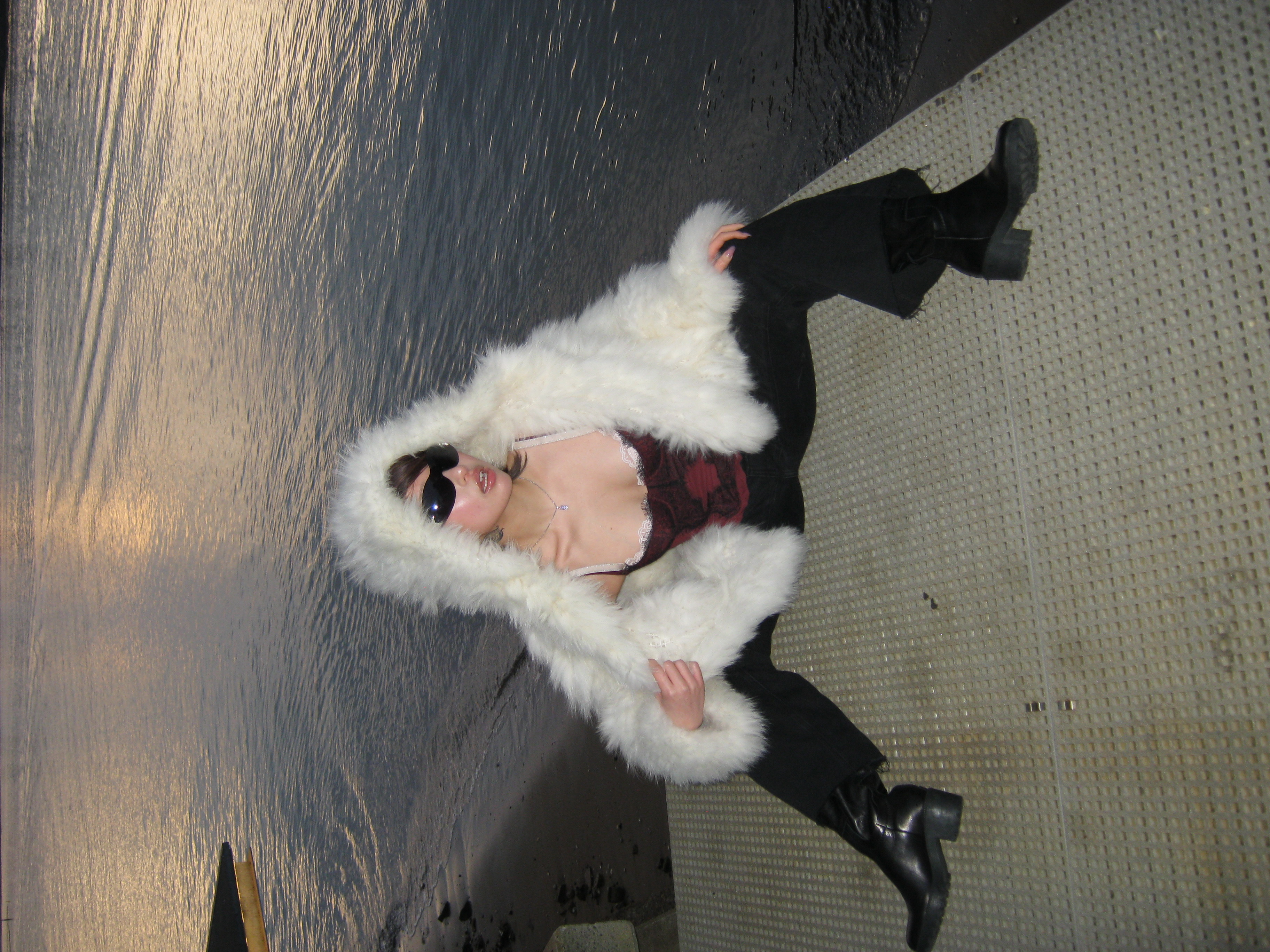 A woman wearing a white faux fur hooded coat and sunglasses, sitting on a dock with water in the background at dusk.