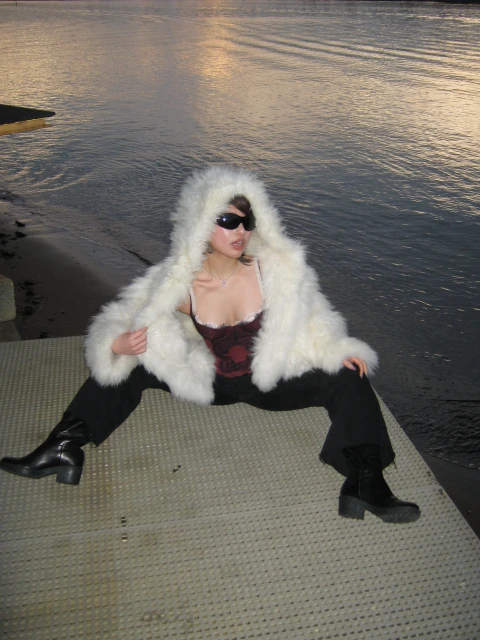 A woman wearing a white faux fur hooded coat and sunglasses, sitting on a dock with water in the background at dusk.
