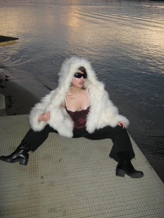 A woman wearing a white faux fur hooded coat and sunglasses, sitting on a dock with water in the background at dusk.
