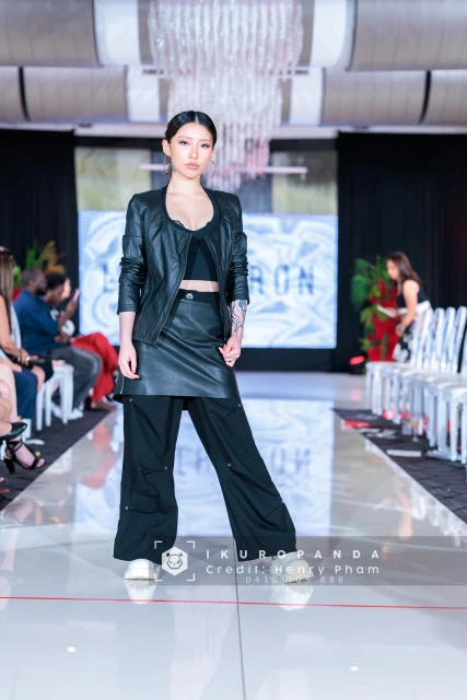 Fashion runway show featuring a woman in a black jacket, bralette, skirt, and pants walking a white runway with seated audience.