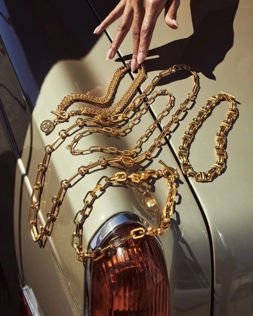 Gold chain necklaces are arranged on the hood of a car, with a hand lightly touching the chains in a fashion editorial setting.