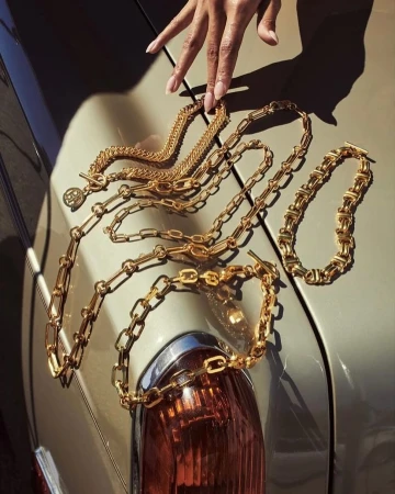 Gold chain necklaces are arranged on the hood of a car, with a hand lightly touching the chains in a fashion editorial setting.