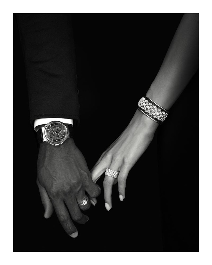 Monochromatic image of two adults holding hands, both wearing diamond jewelry, against a dark background. Wristwatch and suit sleeve visible.