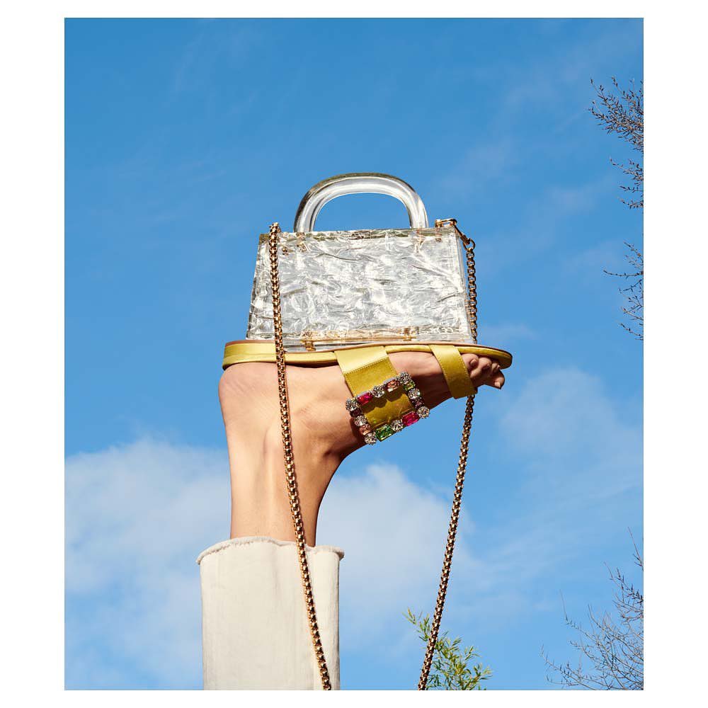 Hand wearing a gold sandal and holding a clear handbag against a vibrant blue sky with wispy clouds and glimpses of trees.