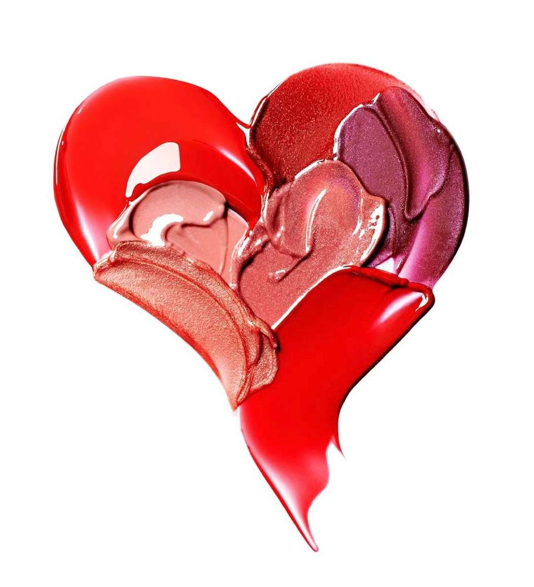 Various shades of red, pink, and coral lip gloss and lipstick swatches create a heart shape on a white background. Beauty and makeup.